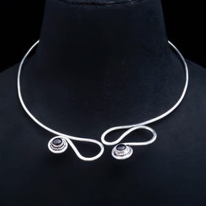 Puede incluir: Un collar tipo gargantilla abierto de plata con dos extremos de alambre asimétricos y en bucle. Cada extremo está acentuado con una pequeña piedra negra redonda y pulida, engastada en un borde decorativo de plata trenzada.