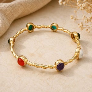 Pode incluir: Uma pulseira tipo bracelete dourada com uma banda metálica texturizada e ondulada, cravejada com seis pedras preciosas cabochão redondas e multicoloridas. As pedras incluem tons de vermelho, roxo, branco, verde e azul-petróleo escuro.