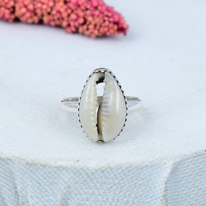 May include: A silver ring featuring a natural, off-white cowrie shell. The shell is oval-shaped with a textured surface and a dark edge. The ring's band is a simple, polished silver. The background is a soft, white surface.