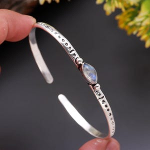 Könnte beinhalten: Ein silbernes Armreif-Armband mit einem ovalen Mondstein in der Mitte. Das Armband hat geätzte Himmelsmotive und kleine dekorative Perlen. Das offene Design ermöglicht ein einfaches Tragen.