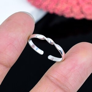 Könnte beinhalten: Ein silberfarbener Zehenring mit einem gedrehten Design. Der offene Ring wird zwischen zwei Fingern vor einem schwarzen Hintergrund gehalten. Das Design des Rings weist eine Reihe von ineinander verschlungenen Kurven auf.
