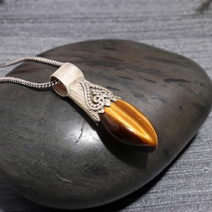 May include: A pendant necklace featuring a polished, oval tiger's eye gemstone. The stone is set in a silver-colored metal with an ornate, decorative design. The necklace chain is a fine, silver-colored link.