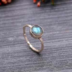 May include: A silver ring with a round, iridescent blue gemstone. The ring has a textured band and a simple, elegant design. The gemstone is set in a bezel setting, and the ring is displayed on a dark gray surface.