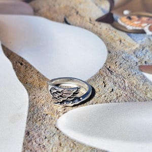 Puede incluir: Un anillo plateado con un diseño de ala. El anillo presenta un motivo de ala detallado en la parte delantera, con la banda curvándose para encontrarse con el ala. El anillo está sobre una superficie texturizada y multicolor.