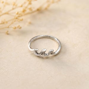 Può includere: Un anello in metallo color argento caratterizzato da un design ondulato continuo sulla parte superiore della fascia. Il metallo presenta una finitura opaca leggermente testurizzata e la fascia mantiene una forma circolare costante.
