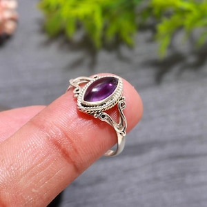 May include: A silver ring featuring a marquise-cut, deep purple gemstone. The gemstone is set in a detailed, ornate silver band with a twisted rope design around the stone. The ring is displayed on a finger.