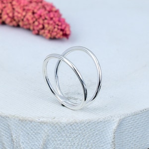 Könnte beinhalten: Ein silberner Ring mit Doppelband-Design. Der Ring hat zwei ineinander verschlungene, glatte Bänder, die eine moderne und minimalistische Ästhetik schaffen. Er liegt auf einer weißen Oberfläche, mit einem verschwommenen rosa Blumenelement im Hintergrund.