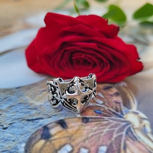 May include: A silver-colored ring with an ornate design featuring a central cross motif and decorative scrollwork. The ring is set against a backdrop of a vibrant red rose and a patterned surface, creating a striking contrast.