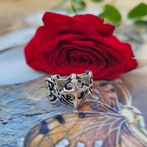 May include: A silver-colored ring with an ornate design featuring a central cross motif and decorative scrollwork. The ring is set against a backdrop of a vibrant red rose and a patterned surface, creating a striking contrast.