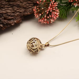 May include: A gold-colored pendant necklace featuring a filigree sphere design. The sphere has intricate cut-out patterns and is suspended from a delicate chain. The necklace is displayed on a white surface with floral accents in the background.