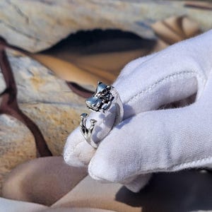 May include: A silver ring with an animal head design. The ring has an open design, with the animal's head on one end and a decorative element on the other. The ring is shown on a white glove. The ring is made of silver.
