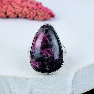 Puede incluir: Un anillo de plata con una piedra preciosa grande en forma de lágrima. La piedra tiene una mezcla de tonos morados oscuros y negros, con una superficie pulida. El anillo está sobre una superficie blanca, con un elemento floral rosa borroso en el fondo.