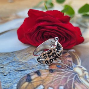 May include: A silver-colored ring with an ornate, filigree design. The ring is set against a backdrop of a vibrant red rose and a patterned surface, creating a striking contrast. The ring's intricate details are highlighted.