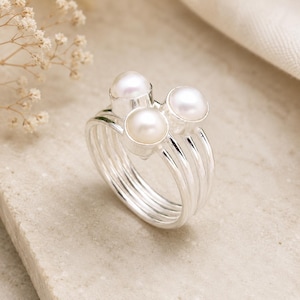 Puede incluir: Un anillo de plata con tres perlas blancas. El anillo tiene un diseño multibanda con un grupo de perlas en la parte superior. Las perlas son redondas y tienen un aspecto brillante. El anillo está sobre un fondo neutro.