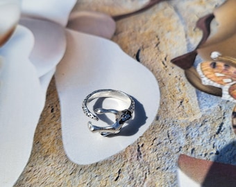 May include: A silver-toned ring with an open design. The band features a textured pattern, and the ring has a unique, abstract shape. The ring is displayed on a light-colored surface, showcasing its intricate details. A piece of jewelry.