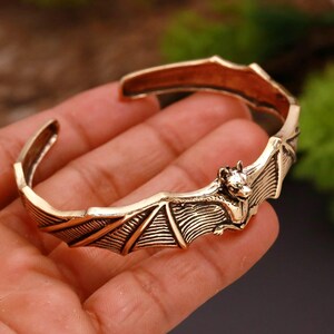 Può includere: Un bracciale a polsino color bronzo con un design a pipistrello. Il pipistrello è centrato sul bracciale, con le ali spiegate. Il bracciale ha una superficie dettagliata e strutturata ed è tenuto in una mano.