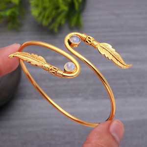 Könnte beinhalten: Ein goldfarbenes Armreif-Armband mit Feder- und Edelsteinakzenten. Das Armband hat zwei kleine, runde Edelsteine und federförmige Designs an jedem Ende. Der Armreif wird von einer Hand gehalten.