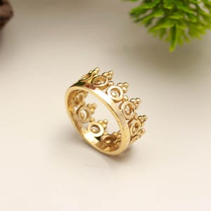 Puede incluir: Un anillo de corona dorado con un diseño detallado. El anillo presenta una serie de elementos circulares y redondeados, creando una apariencia regia. El anillo se encuentra sobre un fondo suave y neutro, resaltando su intrincada artesanía.