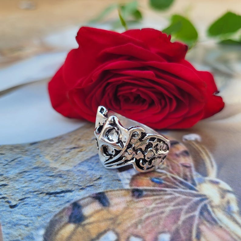 May include: A silver-colored ring with an ornate, openwork design. The ring features a detailed pattern of swirling lines and shapes. A vibrant red rose is in the background.