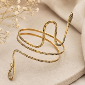 May include: A gold-coloured arm cuff shaped like a snake. The cuff wraps around the arm with a textured, scale-like pattern. The snake's head is detailed with etched features. A unique piece of jewellery.