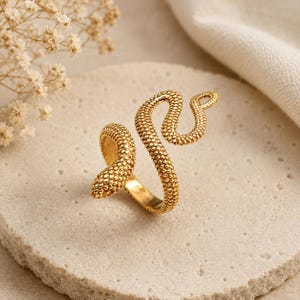 Può includere: Un anello a forma di serpente dorato con una superficie strutturata e puntinata. L'anello si avvolge attorno al dito in un design a spirale, con la testa e la coda del serpente che formano le estremità. L'anello è posto su una superficie di pietra chiara.