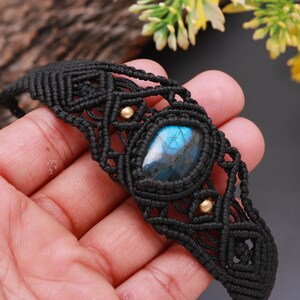 May include: Black macramé bracelet with a central, oval blue gemstone. The bracelet has a woven design with gold accents. The gemstone has a blue and iridescent appearance.