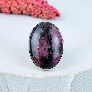 Puede incluir: Anillo ovalado con una piedra preciosa pulida en tonos rosa oscuro y negro, engastada en una banda de metal plateado. El patrón único de la piedra preciosa es la característica principal del anillo.