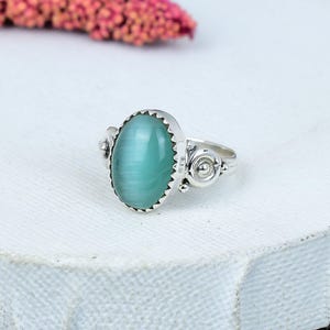 May include: A silver ring featuring an oval, light-green gemstone. The gemstone is set in a scalloped bezel. The ring has decorative details on the sides and a simple band. The ring is displayed on a white surface.