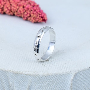 Könnte beinhalten: Ein silberner Ring mit einem Band, das ein Muster aus Sternen und anderen Himmelsdesigns aufweist. Der Ring ist vor einem weißen Hintergrund platziert, mit einem verschwommenen rosa Blumenelement im Hintergrund.