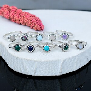 May include: A selection of sterling silver rings, each with a different gemstone. The rings have a sunburst design around the stones, such as amethyst, turquoise, and moonstone. They are displayed on a white surface, with a pink floral arrangement.