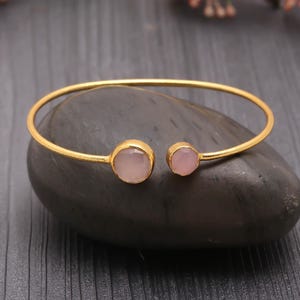Puede incluir: Un brazalete dorado con dos piedras preciosas redondas de color rosa pálido. El brazalete está colocado sobre una piedra gris oscura, con un diseño sencillo y elegante. Las piedras están engastadas en biseles dorados.