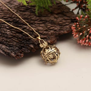 May include: A gold-colored pendant necklace featuring a filigree sphere design. The sphere is intricately detailed with openwork patterns. The necklace chain is delicate and complements the pendant. The background includes a textured wooden surface and greenery.