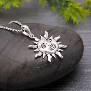 Può includere: Una collana con pendente in argento raffigurante un sole sorridente con raggi. Il pendente è dettagliato con occhi, naso e bocca sorridente. La catena della collana è una catena a cavo argentata e strutturata. Il pendente è esposto su una pietra grigio scuro.