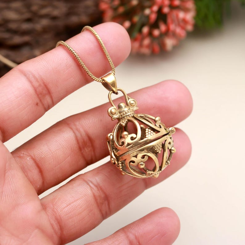 May include: A gold-colored pendant necklace featuring a filigree orb design. The pendant is suspended from a delicate chain. The orb has intricate, openwork patterns and a decorative top. The necklace is held in a hand.