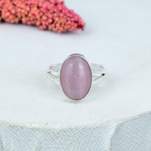 Op de afbeelding: Een zilveren ring met een grote, ovale, lichtroze edelsteen. De ring heeft een gespleten bandontwerp. De edelsteen is glad en gepolijst, gezet in een eenvoudige, elegante zetting. De ring wordt op een wit oppervlak weergegeven.