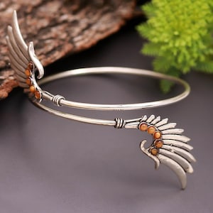 May include: A silver arm cuff with wing-shaped details. The cuff has a curved design with feather-like elements and small, orange-brown gemstones. The jewellery is displayed against a natural wood and green foliage background.