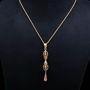 Peut inclure: Un collier de couleur dorée composé d'une fine chaîne et d'un pendentif en forme de vajra, objet rituel aux extrémités symétriques et ornées, se terminant par une pointe lisse et allongée en forme de goutte d'eau.