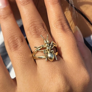May include: A gold-colored ring featuring a detailed ant design. The ring has a double-band design and the ant is sculpted with intricate details, including antennae and legs. The ring is worn on a finger, showcasing the insect-themed jewelry.