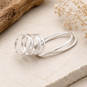 May include: A silver-toned multi-finger ring featuring four circular bands connected to a single elongated oval loop. The piece is crafted from a smooth, polished metal with a continuous design that spans across multiple fingers when worn.
