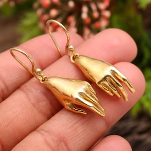May include: A pair of gold-colored earrings, each shaped like a hand with detailed fingers. The earrings have a hook closure and are displayed against a blurred background of greenery.
