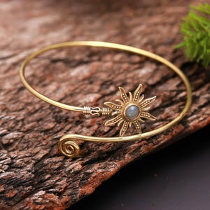 May include: A gold-coloured cuff bracelet with a sunburst design and a central stone. The bracelet has a spiral detail at one end and a decorative clasp. The sunburst has detailed rays and a central, round stone.