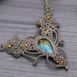 May include: A handmade macrame necklace featuring a teardrop-shaped labradorite stone with blue and green hues. The gray macrame is accented with small gold-colored beads. The necklace is displayed on a gray wooden surface.