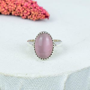 Op de afbeelding: Een zilveren ring met een grote, ovale, lichtroze edelsteen. De edelsteen is gezet in een decoratieve bezel met een geschulpte rand. De ring wordt gepresenteerd op een wit oppervlak, met een vage roze bloemstuk op de achtergrond. Sieraden.