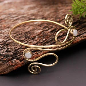 May include: Gold-toned arm cuff with a swirling design and two small, round, white gemstones. The cuff features a hammered texture and is displayed on a piece of wood.
