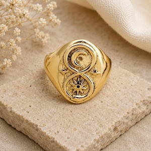Peut inclure: Une bague dorée avec un motif détaillé représentant un croissant de lune et une étoile. La bague est posée sur une surface en pierre claire, mettant en valeur ses détails complexes et sa finition polie.