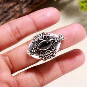 Puede incluir: Colgante ovalado de plata con una piedra preciosa negra en el centro. El colgante presenta intrincados detalles de filigrana y cuentas alrededor de la piedra preciosa. El engaste de plata tiene un acabado pulido, y el colgante se sostiene en una mano.
