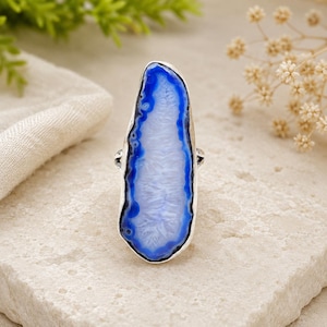 May include: A silver ring with a large, elongated blue and white agate stone. The stone has a striking blue border and a white, crystalline centre. The ring is set on a neutral-toned surface, with greenery and dried flowers in the background.