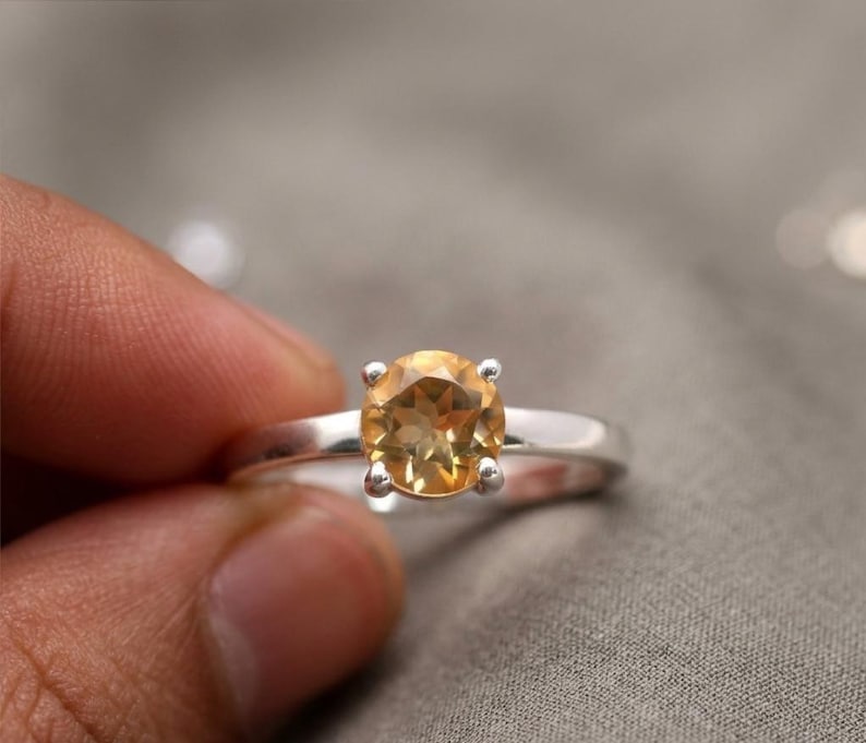 Puede incluir: Un anillo de plata con una piedra preciosa redonda, facetada y de color amarillo dorado, montada en un engaste de cuatro garras. La banda es lisa y pulida, mostrando una piedra clara de tonos c&aacute;lidos que refleja la luz en su superficie.