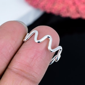 Op de afbeelding: Een zilverkleurige ring met een golvend ontwerp. De ring is een doorlopende, golvende lijn, die een unieke en moderne esthetiek creëert. De ring wordt op een vinger getoond tegen een wazige achtergrond.