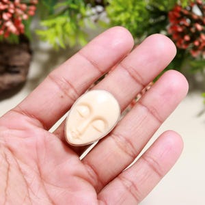 May include: A cream-colored ring with a carved face design. The ring features a silver band and a smooth, oval-shaped face with closed eyes and delicate facial features. The ring is held in a hand.